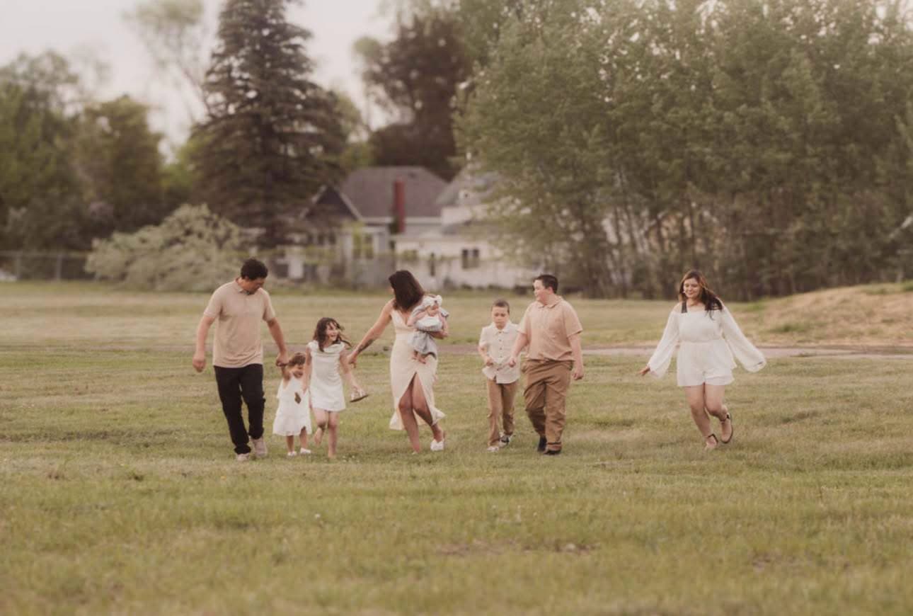 Large family in neutral tones walking and running through a wide, grassy field with trees.
